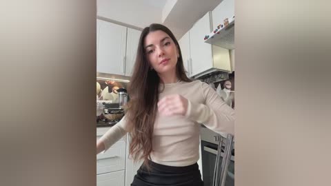 Media: Video of a young woman with long, wavy brown hair, fair skin, wearing a beige long-sleeve top and black pants, standing in a modern, minimalist kitchen with white cabinets and stainless steel appliances.
