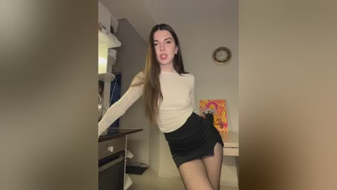 Media: A video of a slender, fair-skinned woman with long, straight brown hair, wearing a tight, cream long-sleeve top and a short, black skirt, posing provocatively in a dimly lit room with beige walls and a colorful painting.