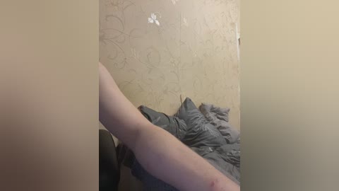 Video of a light-skinned arm, bent at the elbow, reaching into a messy gray bed with rumpled sheets. The background features a beige wall with a floral pattern.