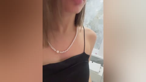 Video of a woman with light skin, straight brown hair, wearing a black spaghetti-strap top and a pearl necklace, standing indoors with blurred background.