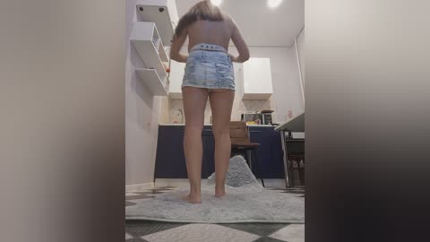 Media: Video of a topless woman with long brown hair, wearing denim shorts, standing barefoot on a fluffy rug in a modern kitchen with white cabinets and a wooden table.