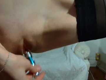A close-up video shows a woman's torso, focusing on her vulva, as she inserts a blue vibrator. The background features a bed with white sheets, a white fan, and a black object. The image is explicit and intimate.