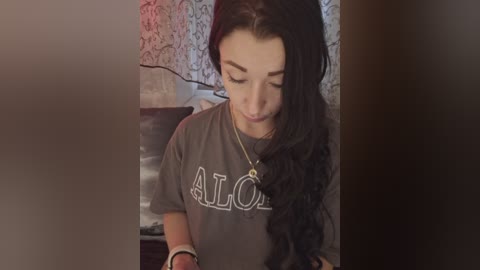 Video of a young woman with long black hair, wearing a gray Aloha shirt, leaning over a table, in a dimly lit room with floral-patterned wallpaper.
