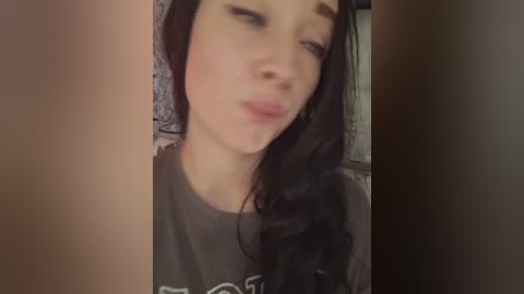 A close-up video of a young woman with long, dark hair, pale skin, and slightly puffy lips, wearing a gray T-shirt. The background is blurred and dimly lit.