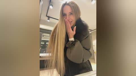 Video of a blonde woman with long hair, wearing a grey puffer jacket, holding her hair, smiling in a modern, well-lit salon with light wood and grey accents.