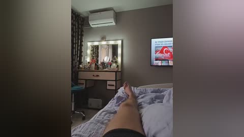 Media: Video of a dimly lit bedroom with a person's legs and feet visible in the foreground, resting on a bed with grey sheets. A wall-mounted television displays a news program, and a vanity table with a mirror and makeup items is visible in the background.