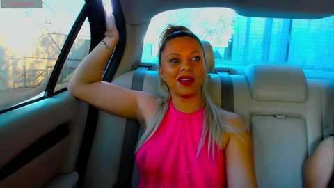 Media: Video of a blonde woman with red lipstick, wearing a sleeveless pink top, sitting in a car with a blue tinted window.