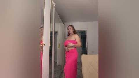 Media: Video of a curvy, fair-skinned woman with long brown hair in a bright pink strapless dress, standing in a modern, minimalistic living room with white walls, a beige chair, and a mirrored closet.