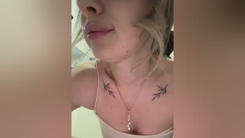 A video of a fair-skinned woman with blonde hair, wearing a beige camisole, showcasing two small bird tattoos on her collarbone. She has a delicate necklace. The background is blurred.