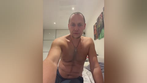 Media: Video of a shirtless, muscular man with short hair, wearing jeans, standing in a modern bedroom with beige walls, a bed, and abstract art on the wall.