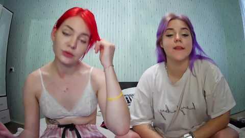 Media: Video of two young women with red and purple hair, wearing white lace bras and t-shirts, sitting on a bed in a bedroom with light blue wallpaper.