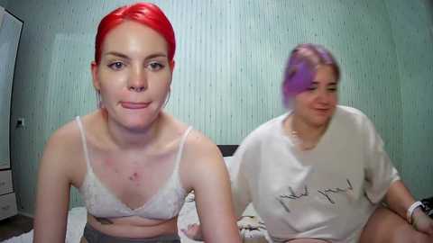 Media: Video of two young women with fair skin and red and purple hair, wearing white lace bras and loose-fitting shirts, sitting on a bed in a dimly lit room with pale green textured wallpaper.