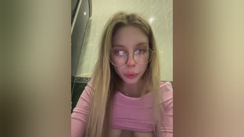 Video of a young woman with fair skin and long blonde hair, wearing glasses and a tight pink crop top, puckering her lips, in a bathroom with beige tiled walls and a shower curtain.