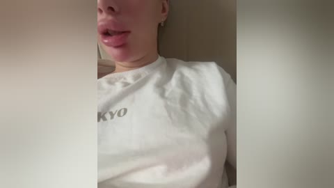 Video of a Caucasian woman with fair skin, wearing a white t-shirt with \"KYOTO\" in black text, and sticking her tongue out playfully. The background is blurred, showing a beige wall.