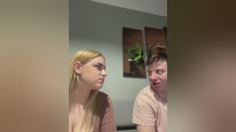 Video of a blonde woman and a man with short brown hair, both making funny faces, in a room with gray walls and abstract art.