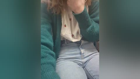 Video of a young person with curly hair, wearing a teal cardigan over a white blouse and light blue jeans, sitting with head in hands, blurred background.