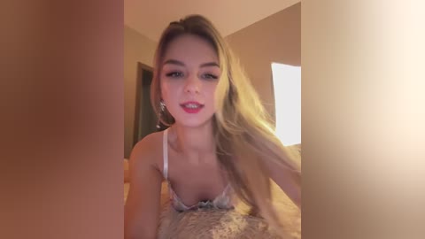 A video of a young, blonde Caucasian woman with fair skin, wearing a white lace bra, lying on a bed with beige sheets, in a softly lit bedroom.