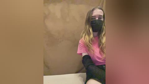 A video of a young woman with long blonde hair, wearing a black face mask, pink t-shirt, black shorts, and black elbow-length gloves, sitting in a dimly lit room with brown walls and a beige floor.