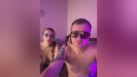 A video of a topless woman with medium-sized breasts and a tattooed arm, wearing glasses and standing beside a topless man with glasses and a tattoo. Both are in a dimly lit room with purple lighting.