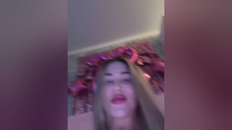 A blurry video of a woman with fair skin and long, wavy hair, wearing a red headband and red lipstick, seen from a low angle, with a pinkish hue dominating the background.
