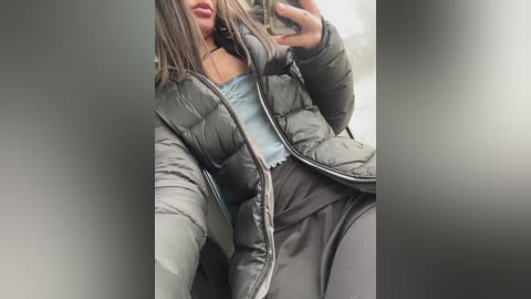 Media: Video of a young woman with light brown skin, wearing a gray puffy jacket, black leggings, and a blue top, taking a selfie in a dimly lit room.