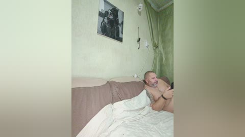 Video of a bearded, nude man lying on a bed with white sheets, in a small, dimly lit room with green curtains and a black-and-white photo on the wall.