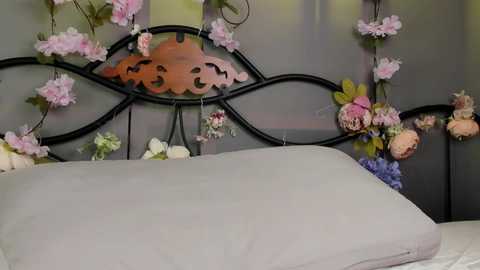 A video of a bed with a black wrought iron headboard adorned with pink cherry blossoms, floral garlands, and a carved wooden pumpkin. The bed is covered with a light gray duvet and pillow, set against a muted gray wall.