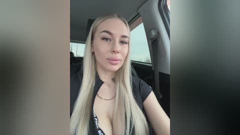Video of a light-skinned, blonde-haired woman with long hair, wearing a black top and necklace, sitting in a car.