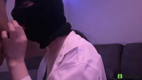 A video of a person with a black hood and white shirt, partially unbuttoned, covering their mouth, standing in a dimly lit room with a gray couch.