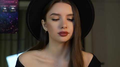 Media: Video of a fair-skinned woman with long brown hair wearing a black hat and black off-shoulder top, set against a blurred, dark background.