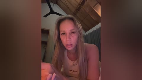 Media: A video of a blonde woman in a white tank top, leaning forward, with a ceiling fan above. The background includes wooden beams and a window with closed curtains.