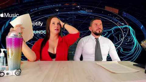 Video of a woman in a red blazer and a man in a white shirt and tie, both seated at a table in a dimly-lit studio with a futuristic, sci-fi backdrop.