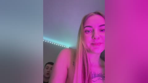 Media: Video of a blonde woman with fair skin, wearing a pink bra, standing in a room with pink and blue lighting. Behind her, a man with short dark hair and a tattoo is visible.