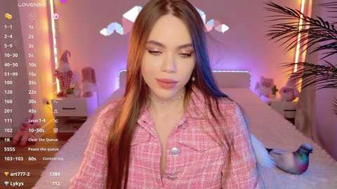 Media: Video of a young woman with long, straight, brown hair, wearing a pink plaid shirt, sitting in a cozy, pink-lit bedroom with plush bedding and potted plants.
