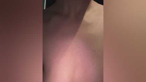 Video of a close-up, side view of a nude person with light skin, showing a slight redness and a small mole on the right shoulder. The background is blurred and dimly lit, highlighting the subject's body.