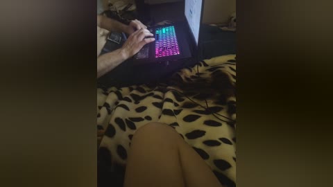 A video of a person's legs, wearing black shorts, lying on a bed with a leopard-print blanket. In the background, a person is typing on a glowing keyboard, with their hands visible. The room is dimly lit, creating a cozy, intimate atmosphere.