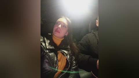 Video of a woman with long brown hair, wearing a yellow turtleneck under a black leather jacket, sitting in a dark car.