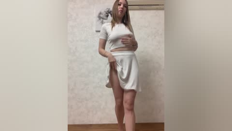 A video of a young Caucasian woman with light skin and shoulder-length blonde hair, wearing a white crop top and skirt, standing against a textured, off-white wall in a plain room.