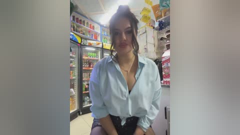 Media: Video of a woman with light skin, dark hair in a ponytail, wearing a light blue blouse, sitting in a brightly lit convenience store with shelves of drinks and snacks in the background.