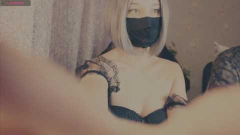 Video of a young woman with light skin, short platinum blonde hair, and a black face mask, wearing a black strapless bra. Her right shoulder has a tattoo. She is indoors with a blurred background.