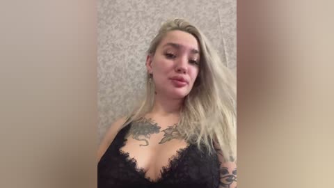 Video of a fair-skinned, blonde woman with long hair, wearing a black lace bra revealing a large dragon tattoo on her chest. Background is a textured beige wall.