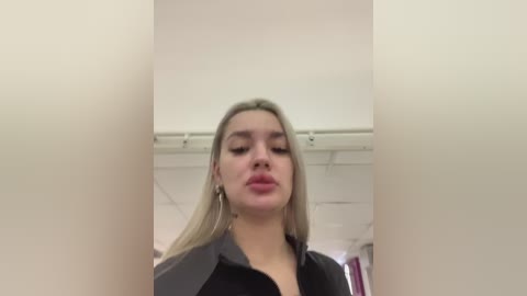 Video of a young, fair-skinned woman with straight, blonde hair, wearing a black blouse, standing in a beige-walled bathroom.