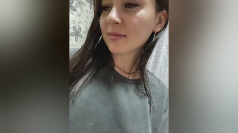 Media: Video of a young woman with long dark hair, fair skin, and light makeup, wearing a grey t-shirt, leaning against a towel in a bathroom with floral wallpaper.