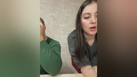 Media: Video of a woman with long, straight dark hair, wearing a black leather jacket, leaning over a table, looking at a man with short hair in a green shirt, in a room with a beige wall.