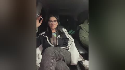 Media: Video of a woman with long black hair and glasses, wearing a black jacket and gray jeans, smoking in a dimly-lit car interior.