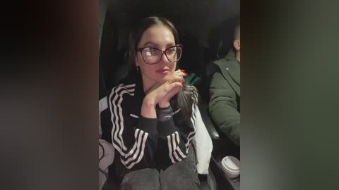 Media: Video of a young woman with glasses, wearing a black Adidas jacket, resting her chin on her hand, seated in a dimly lit car.