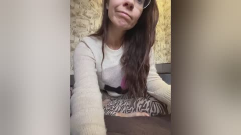 Media: A video of a smiling, fair-skinned woman with long dark hair, wearing glasses, a beige sweater, and zebra-patterned pants, seated against a textured yellow wall.