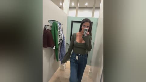 Media: Video of a woman with long brown hair, wearing a green top and high-waisted jeans, taking a selfie in a brightly lit, modern clothing store.