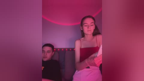 Media: Video of two young people in a dimly lit, red-lit room. A young woman with long brown hair, wearing a red top and white skirt, sits on a black chair. Beside her, a young man with short black hair, dressed in a black t-shirt, gazes at her.