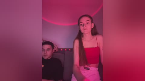 Media: Video of a young man with short dark hair, wearing a black hoodie, and a young woman with long brown hair in a red crop top, standing against a red-lit wall in a dimly lit room.